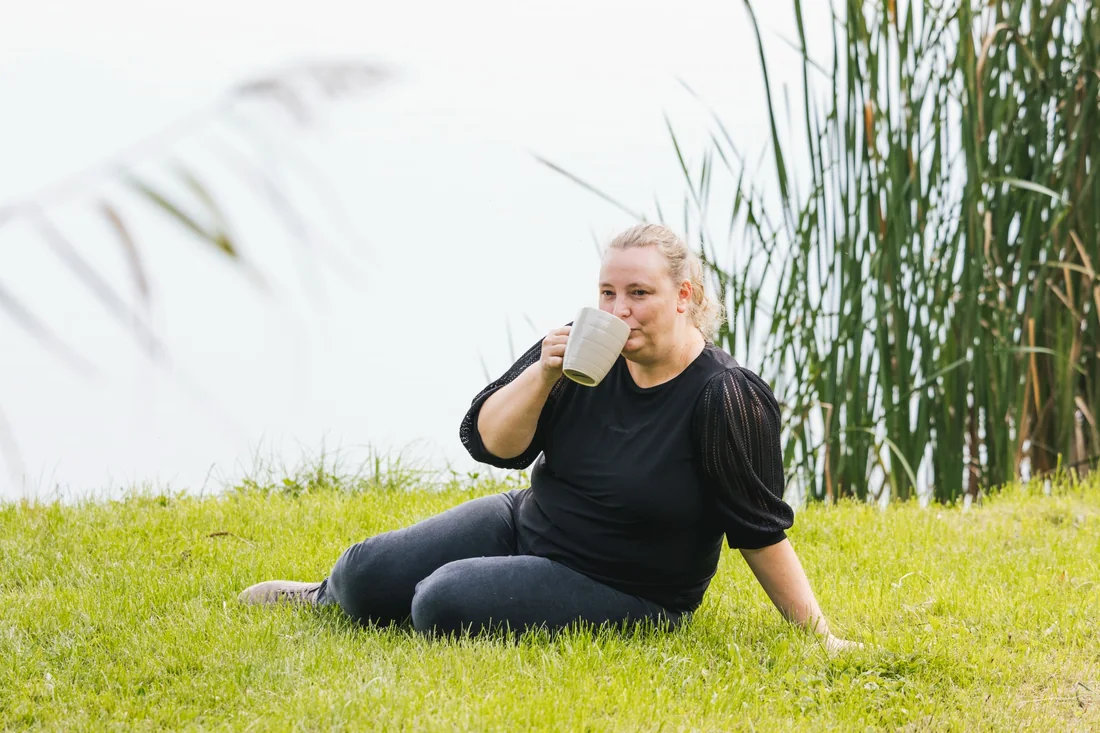 Lindsy zit met een mok in het gras naast het water.